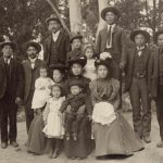 Japanese Family photo with 14 people dressed up with men in suit and ties and women in long dresses. Park setting.