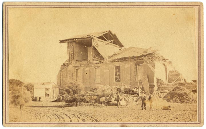 View of collapsed building. Two figures in front beside a pile of logs and brush