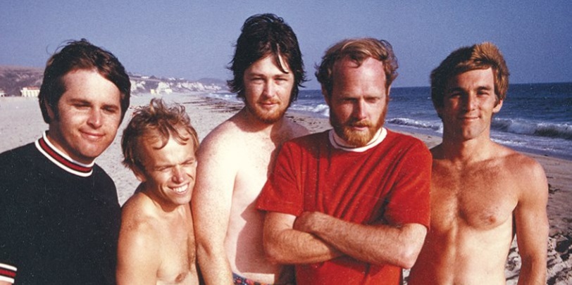 Photo of five smiling young men standing on beach on a breezy, sunny day.