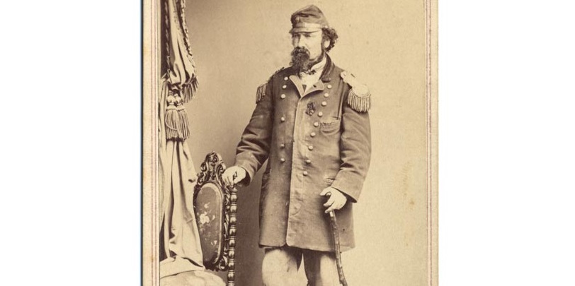 Bearded man in shabby military coat with cap and wooden cane. Standing by a chair