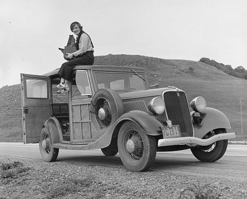 Dorothea Lange on top of old Ford with bare hills behind