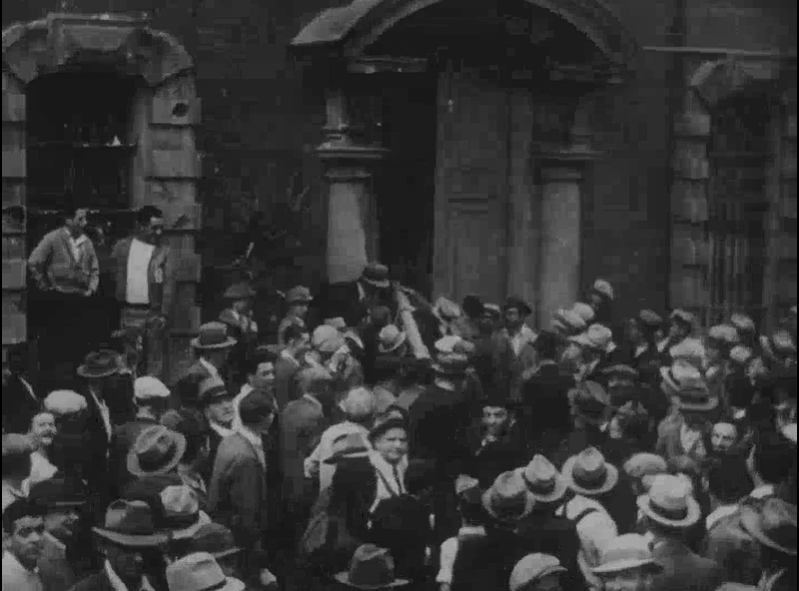 A grayscale image of a group of people, many wearing hats, using a log to batter down a door.