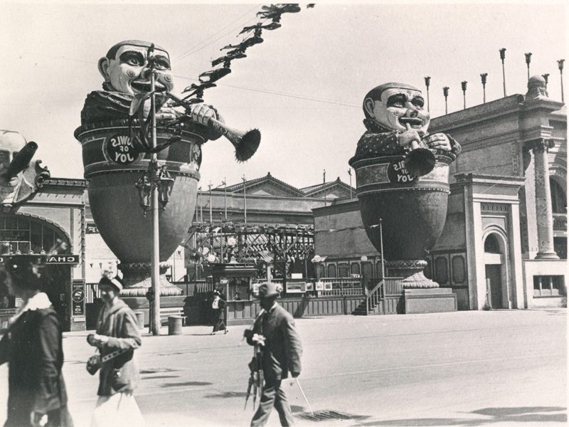 Two large figures of Tweedle Dee and Tweedle Dum, each standing in a bowl and playing a horn, are placed at the entrance to a roller coaster. People stroll by in the foreground.