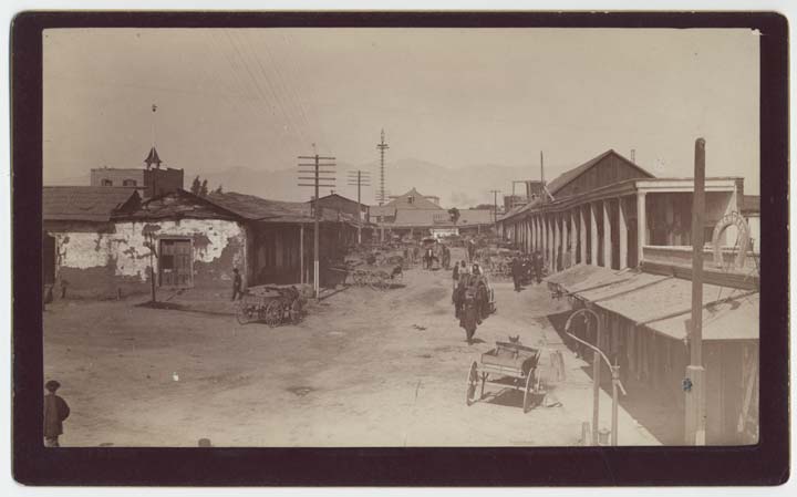 Street scene in Chinese Quarter; shows adobe and frameboard buildings (right and left), carts and wagons (center), people walking (center and at left