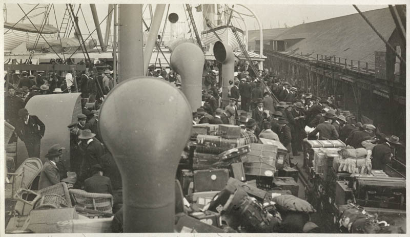 View of many Japanese Immigrants and luggage crowded on the deck of the S.S. Manchuria. Building is seen at right.