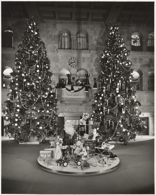 Two three floor high decorated christmas trees stand in front of entrance in lobby with floor display featuring childrens toys on circular platform. At Barker Bros. Photograph dated Dec. 11, 1958.