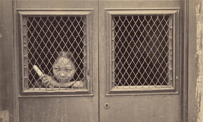 View of a young woman looking out a wire mesh window in a Chinese brothel door.