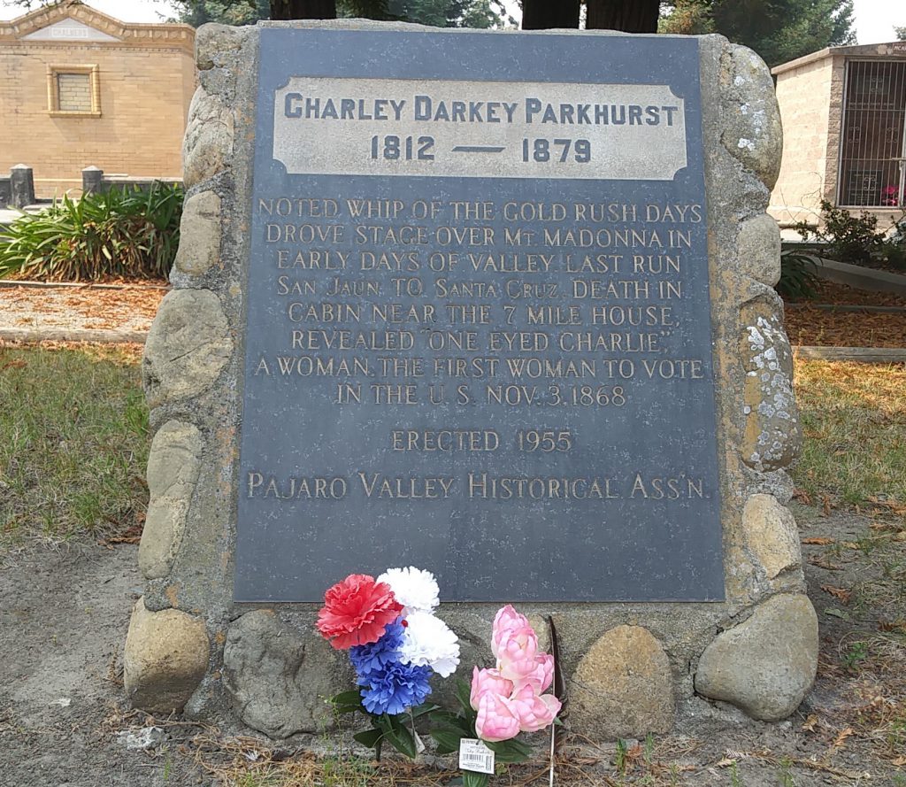 A grave marker made of small stones cemented together surrounding a black plaque. Grave stones and tombs sit on the grass behind. Red, white, and blue flowers have been placed in front of the grave.