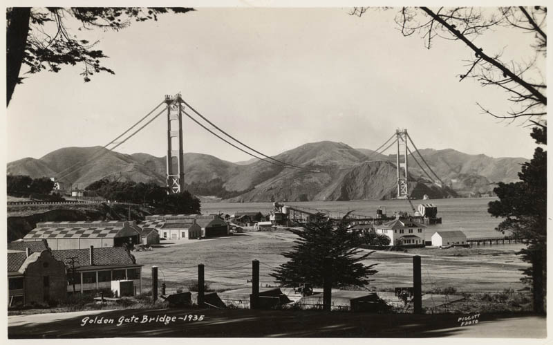 View of the Golden Gate Bridge under construction with the Presidio seen in the foreground and the Marin Headlands in the distance. The bridge towers and suspension cables are seen.