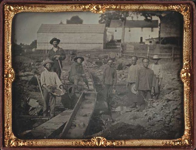 View of seven miners standing next to a sluice box; three white miners standing to the left of the sluice, four Chinese miners standing on the right.