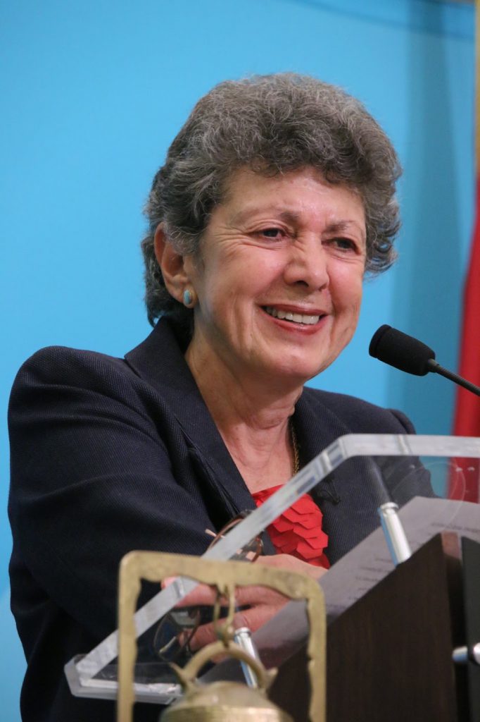 A photograph of a smiling woman with short graying hair wearing a black blazer, standing at a podium.