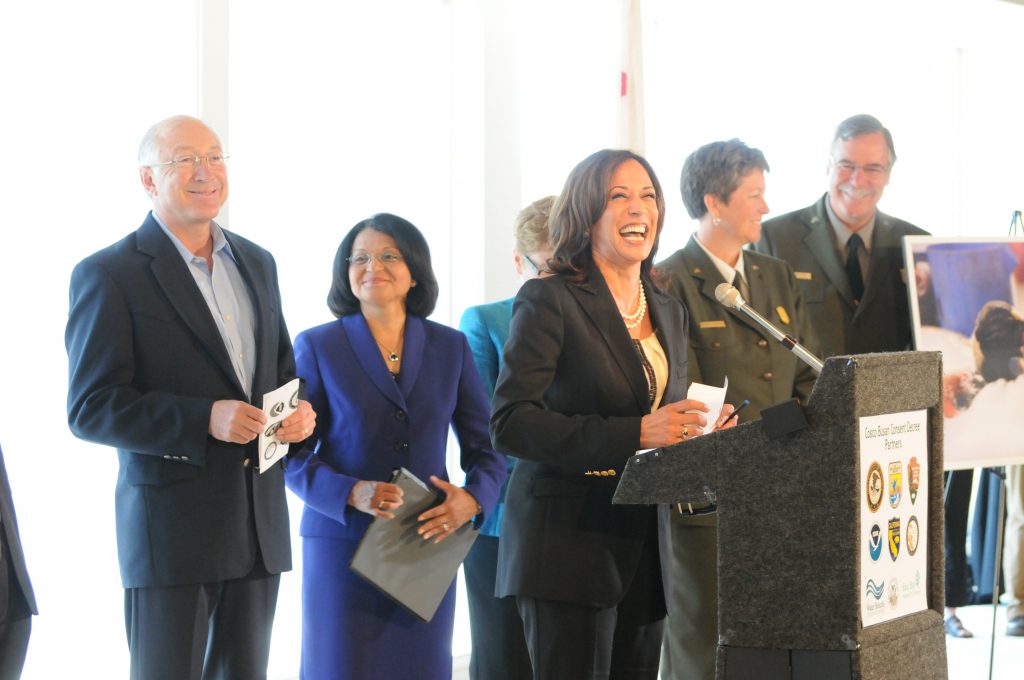 Shows Kamala Harris giving a speech while working as California's Attorney General. Several people including Secretary Ken Salazar are standing behind her.